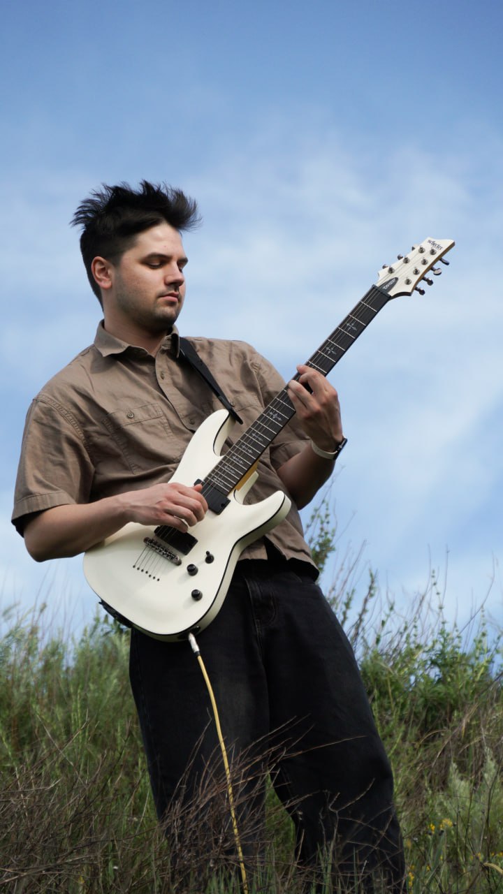Thaddeus playing a white electric guitar on a hillside against an open sky